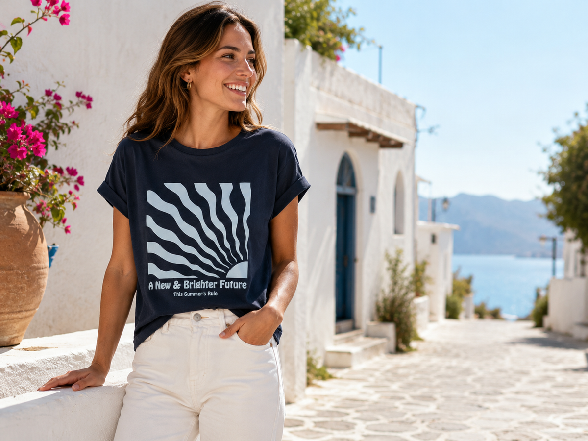 Mujer con camiseta azul marino estampada y pantalón blanco en un entorno mediterráneo junto al mar.